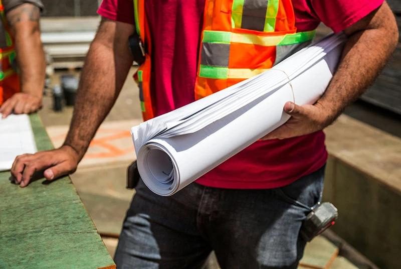 Construction worker in high-visibility vest holding rolled building plans on a construction site.